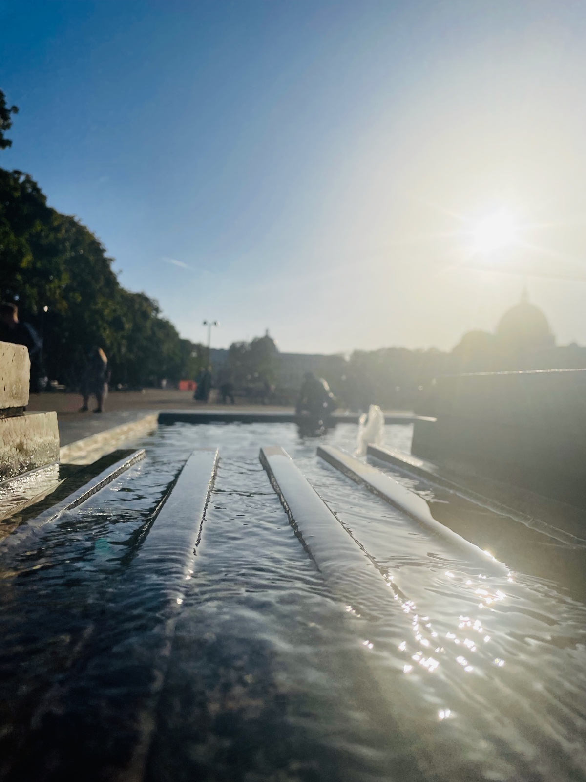 Water flowing down a fountain channel toward a sunlit plaza and distant dome